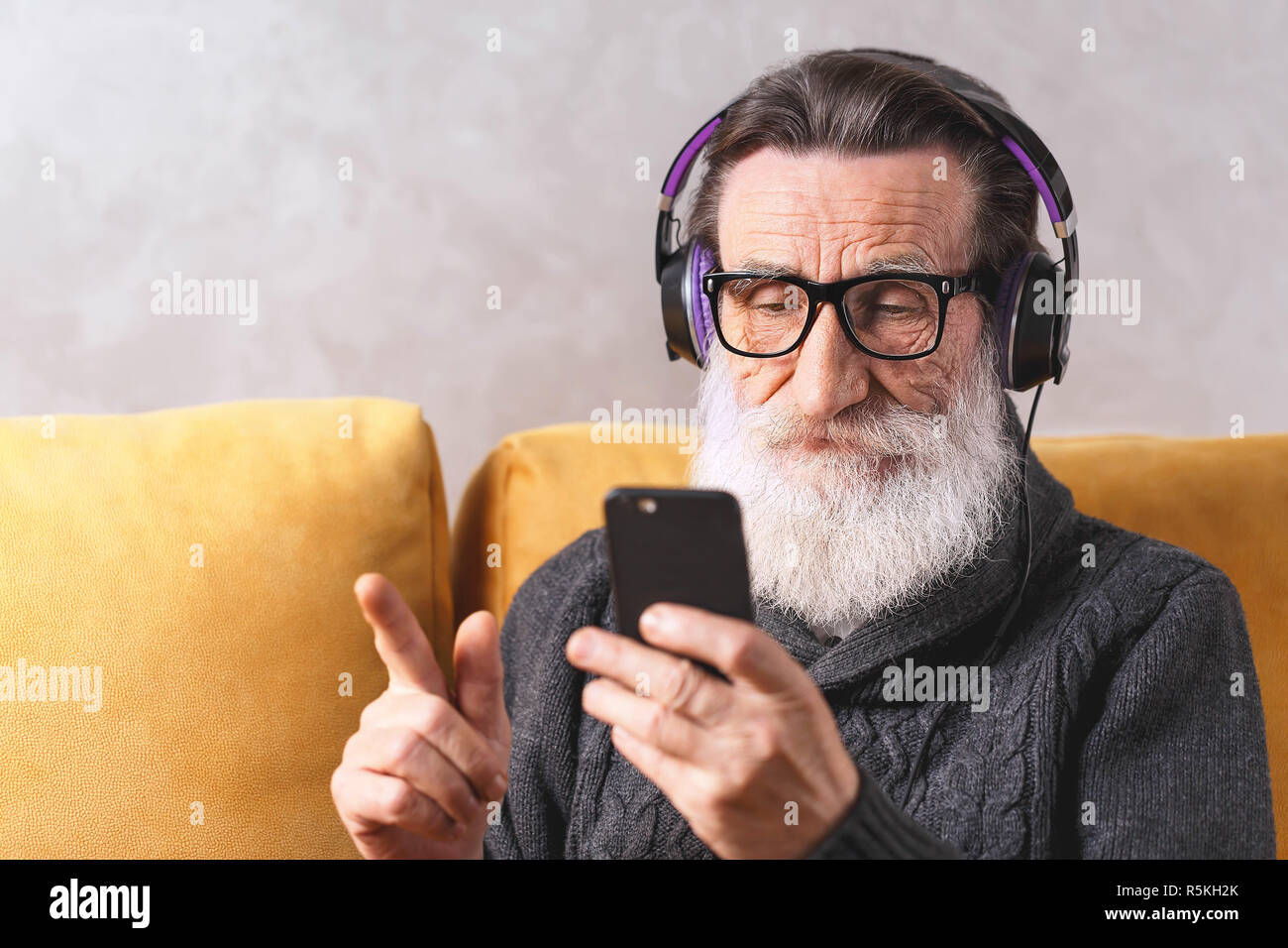 Senior zeitgenössische bärtigen Mann mit Brille grau Pullover lernen Sie das Smartphone während der Sitzung in der Kopfhörer auf ein gelbes Sofa in seinem Lichte Wohnzimmer setzen, moderne Technik, Kommunikation Konzept Stockfoto