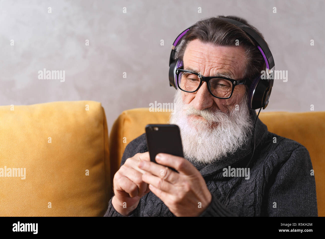 Senior zeitgenössische bärtigen Mann mit Brille grau Pullover writting Nachricht auf dem Smartphone während der Sitzung in der Kopfhörer auf ein gelbes Sofa in seinem Lichte Wohnzimmer, moderne Technologie, Kommunikation Konzept Stockfoto