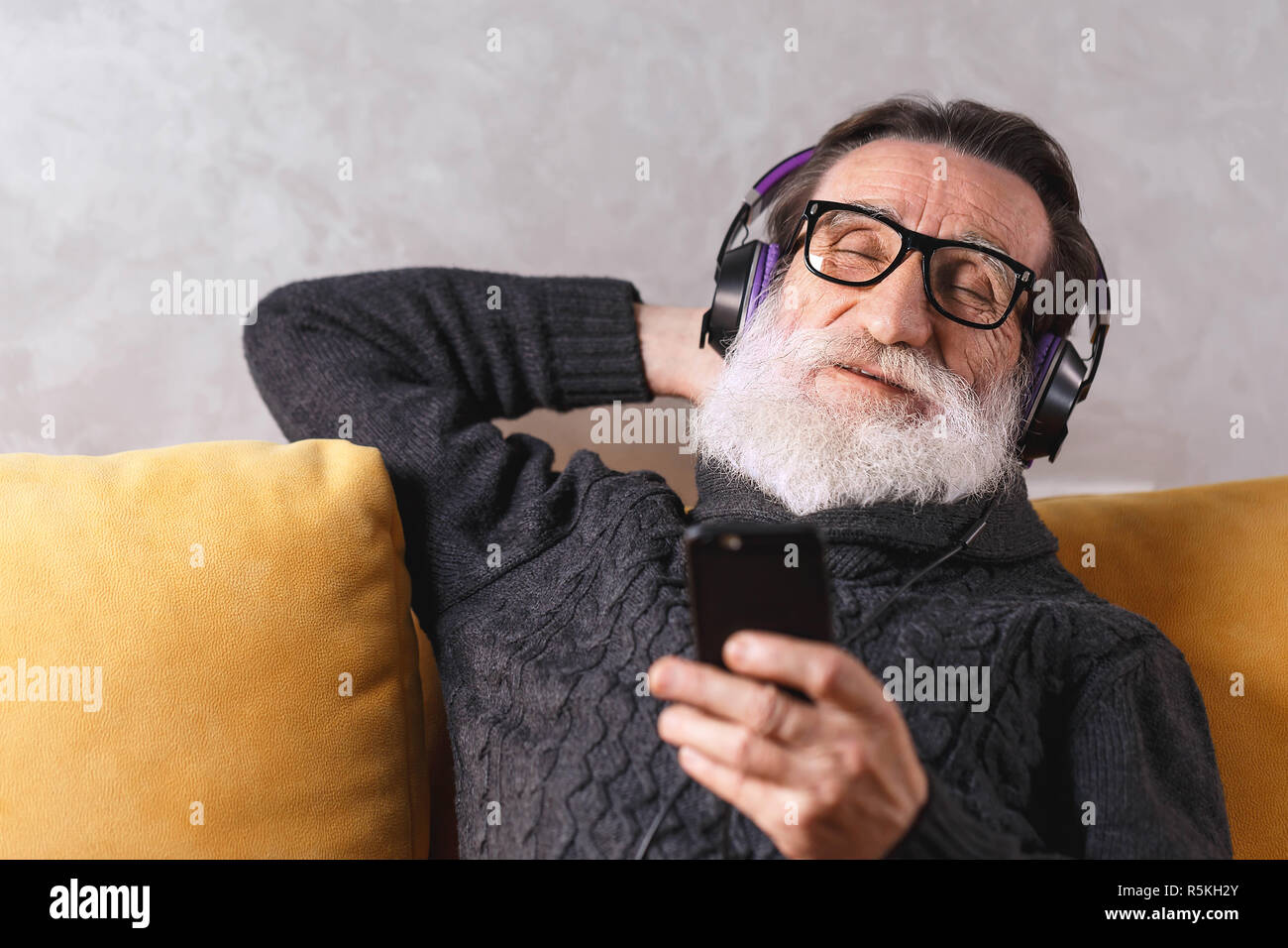 Senior heiter bärtigen Mann mit Brille grau Pullover entspannen mit dem Smartphone während der Sitzung in der Kopfhörer auf ein gelbes Sofa in seinem Lichte Wohnzimmer, er seine Augen schließen und zuhören ein berühmter Musiktitel, moderne Technik, Kommunikation Konzept Stockfoto