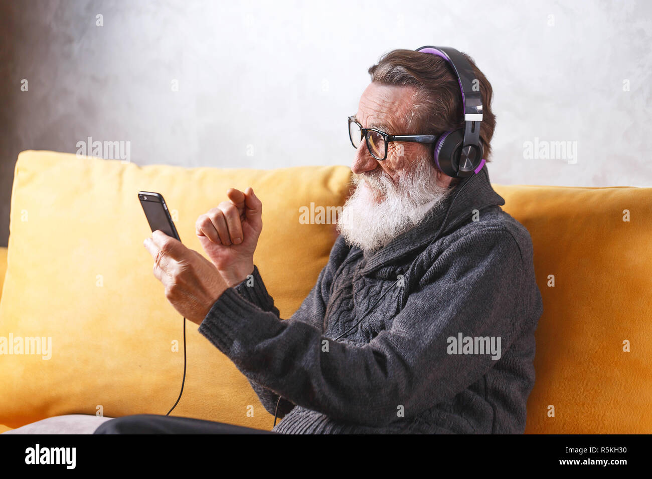 Senior heiter bärtigen Mann mit Brille grau Pullover Sie Ihr Smartphone während der Sitzung in der Kopfhörer auf ein gelbes Sofa in seinem Lichte Wohnzimmer, moderne Technologie, Kommunikation Konzept Stockfoto