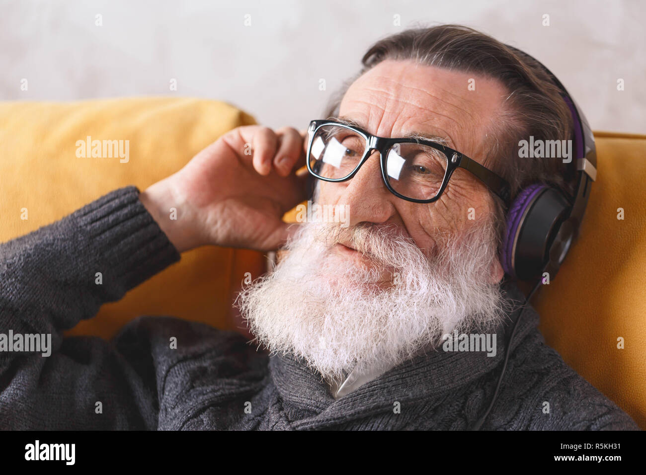 Senior aussehenden bärtigen Mann mit Brille grau Pullover sitzen in der Kopfhörer auf ein gelbes Sofa in seinem Lichte Wohnzimmer und hören seine Lieblingsmusik, moderne Technik, Kommunikation Konzept Stockfoto