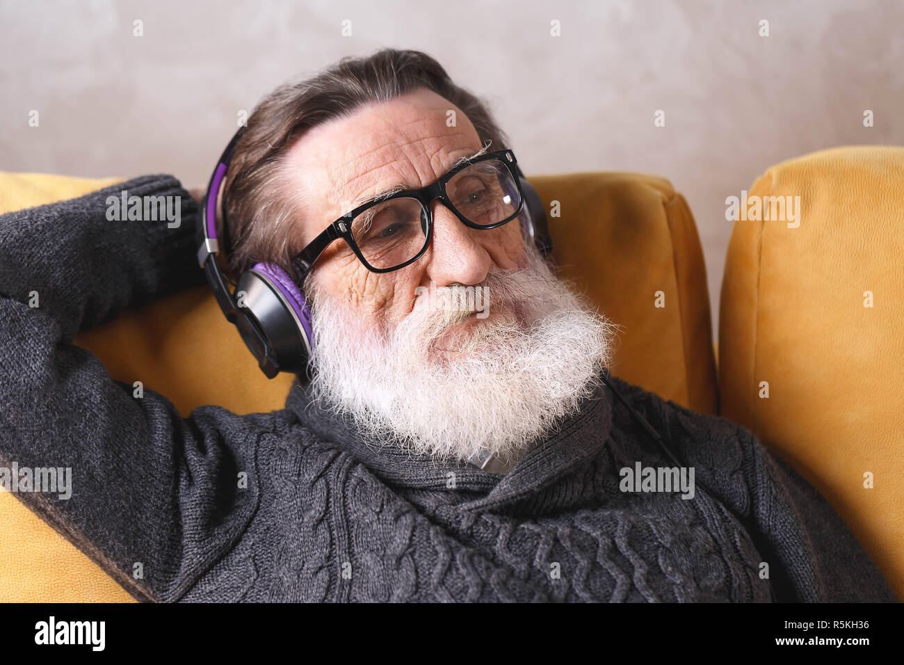 Senior aussehenden bärtigen Mann mit Brille grau Pullover sitzen in der Kopfhörer auf ein gelbes Sofa in seinem Lichte Wohnzimmer, entspannend und hören seine Lieblingsmusik, moderne Technik, Kommunikation Konzept Stockfoto