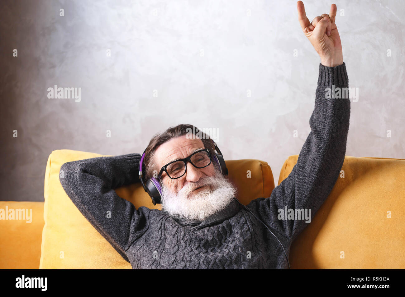 Senior aussehenden bärtigen Mann mit Brille grau Pullover sitzen in der Kopfhörer auf ein gelbes Sofa in seinem Lichte Wohnzimmer, entspannend und hören seine Lieblings rock music Song, moderne Technik, Kommunikation Konzept Stockfoto