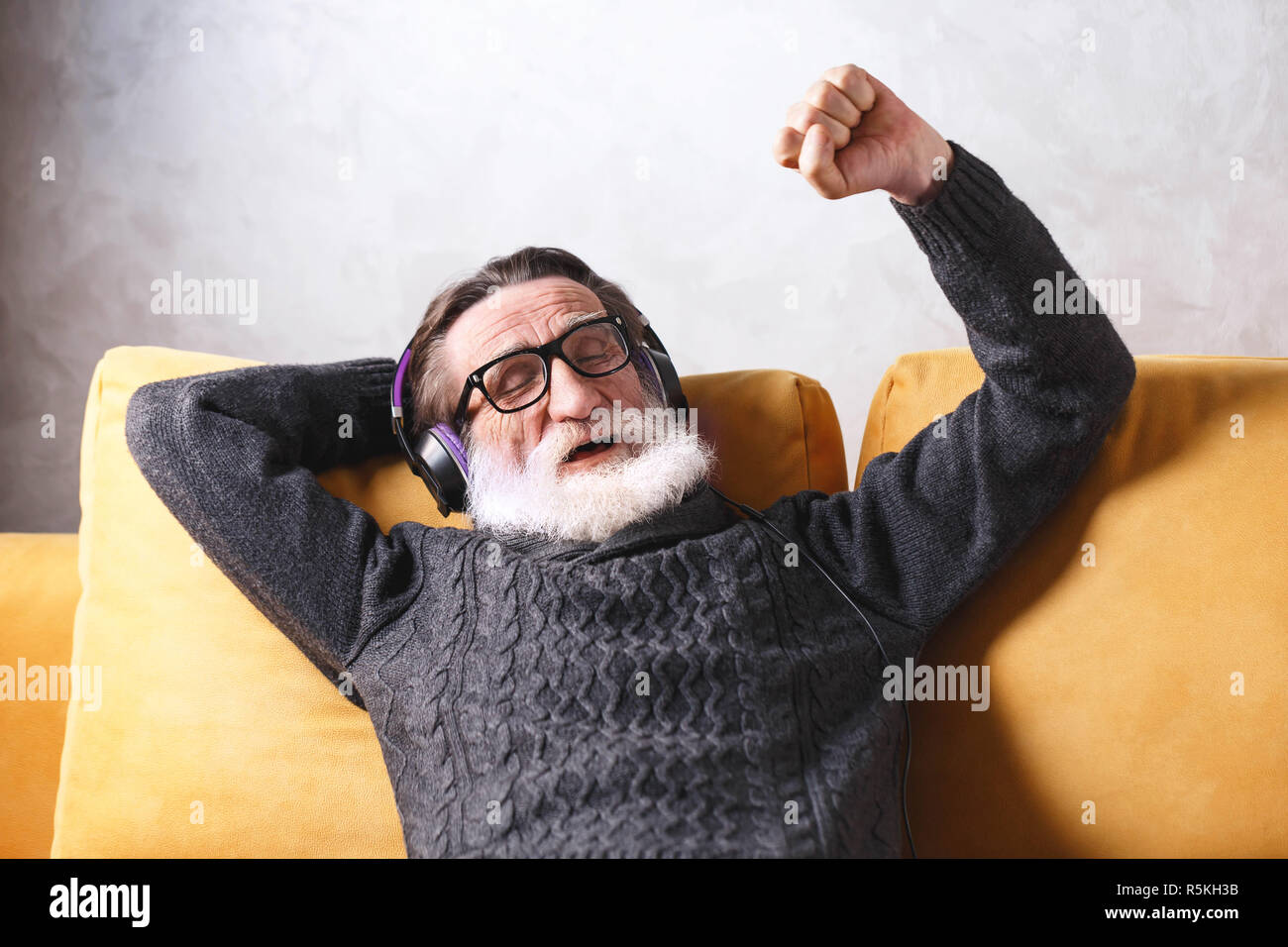 Senior aussehenden bärtigen Mann mit Brille grau Pullover sitzen in der Kopfhörer auf ein gelbes Sofa in seinem Lichte Wohnzimmer, entspannend und hören seine Lieblings rock music Song, er seine Augen schließen und sich in den Song, moderne Technik, Kommunikation Konzept Stockfoto