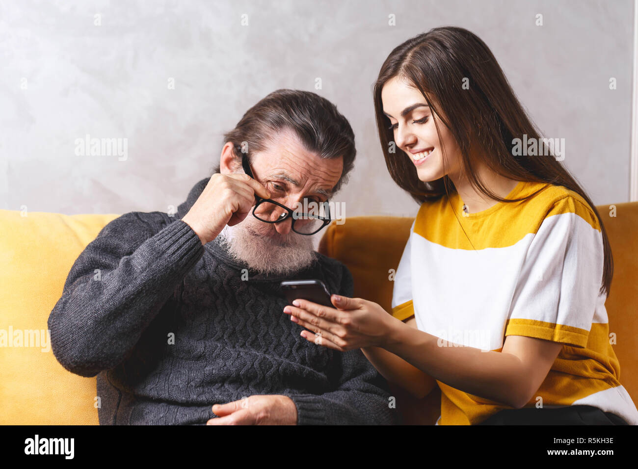 Senior aussehenden bärtigen Mann mit Brille grau Pullover sitzend mit Seiner erwachsenen Langhaarige schöne Tochter auf dem gelben Sofa und ernsthaft auf der Suche auf dem Smartphone, moderne Technik, Kommunikation Konzept Stockfoto
