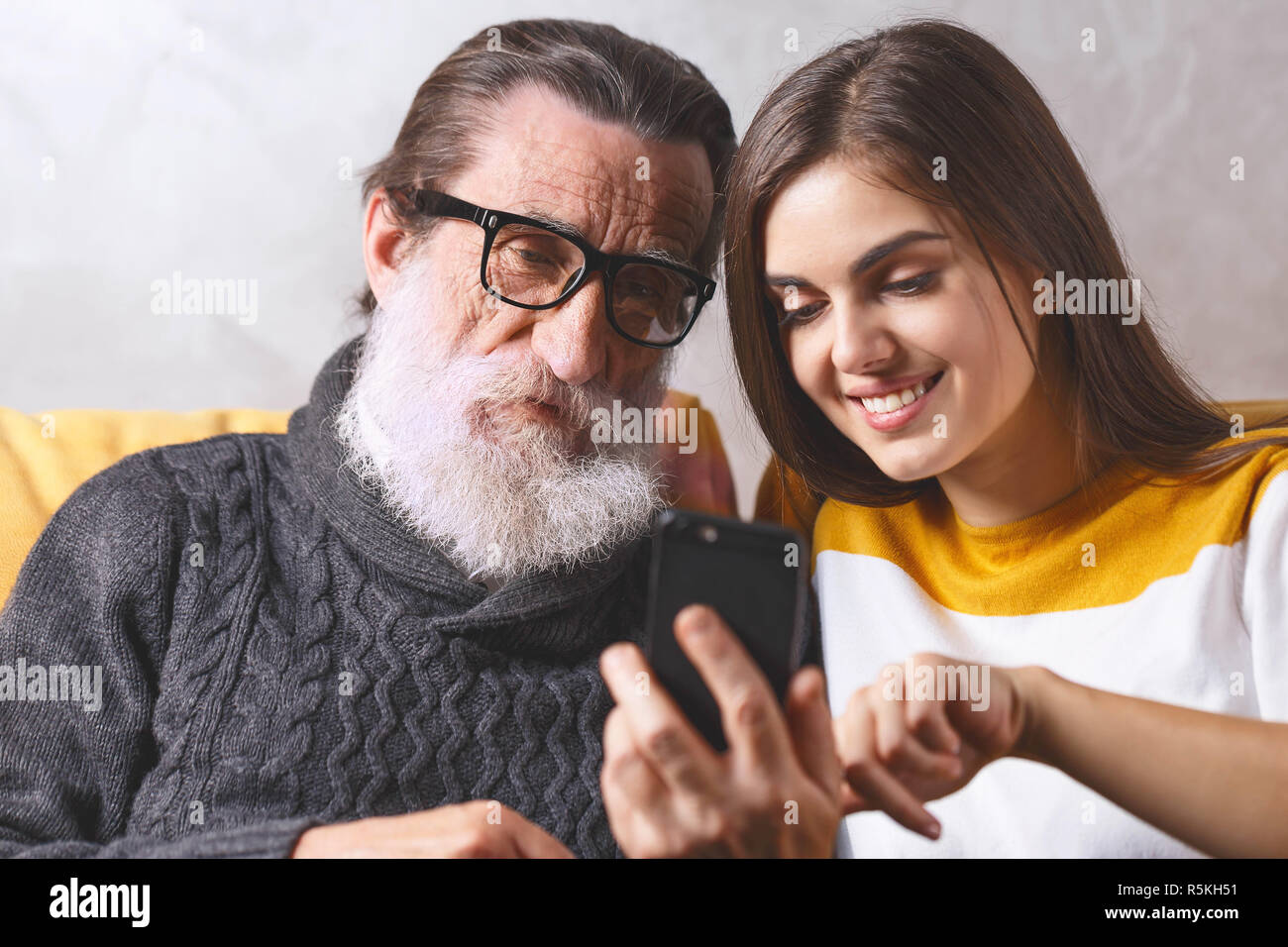 Portrait von älteren bärtigen Vater in Gläsern in Anbetracht der Smartphone beim Sitzen auf dem gelben Sofa im hellen Wohnzimmer mit seinem erwachsenen Langhaarige brünette Tochter, moderne Technik, Kommunikation Konzept Stockfoto