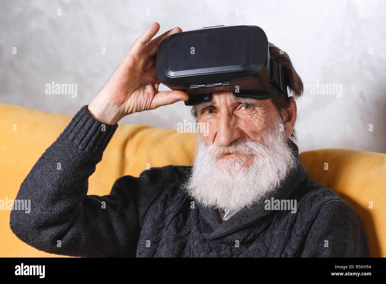 Zeitgenössische älterer bärtiger Mann mit grauen pullover sitzt im VR-Brille auf dem gelben Sofa im Wohnzimmer, er schaut in die Kamera, Technik der Zukunft Konzept Stockfoto