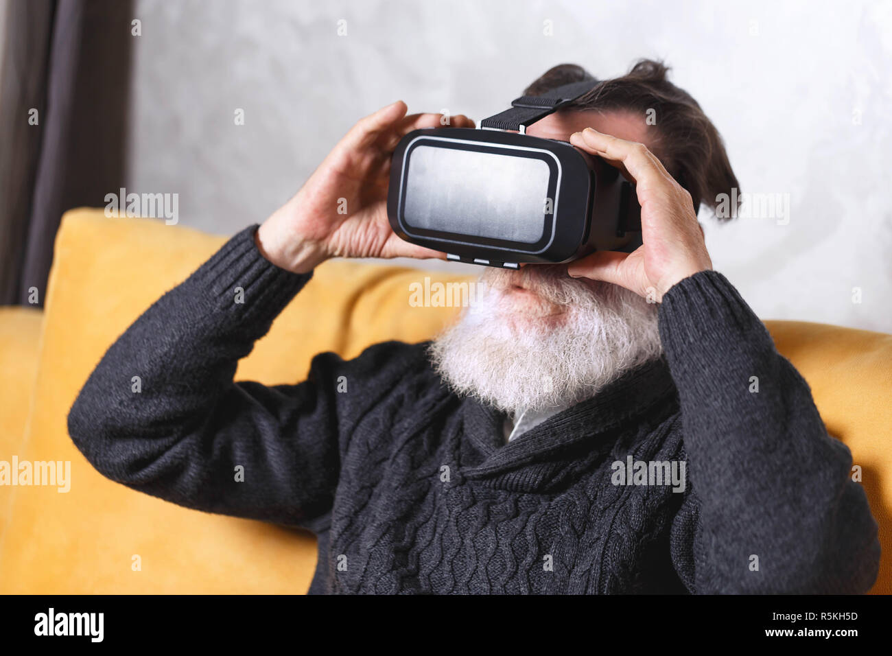 Zeitgenössische älterer bärtiger Mann im grauen pullover Tragen einer Virtual reality Headset beim Sitzen auf dem gelben Sofa im Wohnzimmer, zukünftige Technologie Konzept Stockfoto