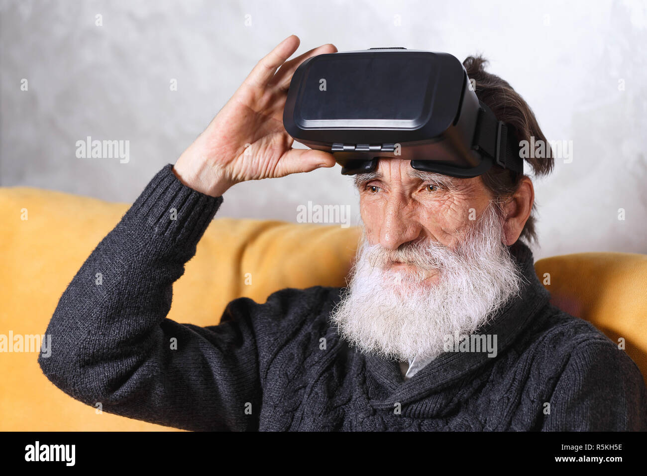 Zeitgenössische älterer bärtiger Mann im grauen pullover sitzt im VR-Brille auf dem gelben Sofa im Wohnzimmer, zukünftige Technologie Konzept Stockfoto