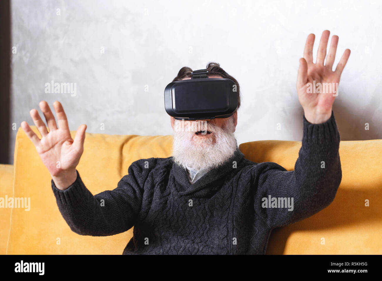 Zeitgenössische älterer bärtiger Mann im grauen pullover Prüfung VR-Gerät während der Sitzung auf dem gelben Sofa im Wohnzimmer, zukünftige Technologie Konzept Stockfoto