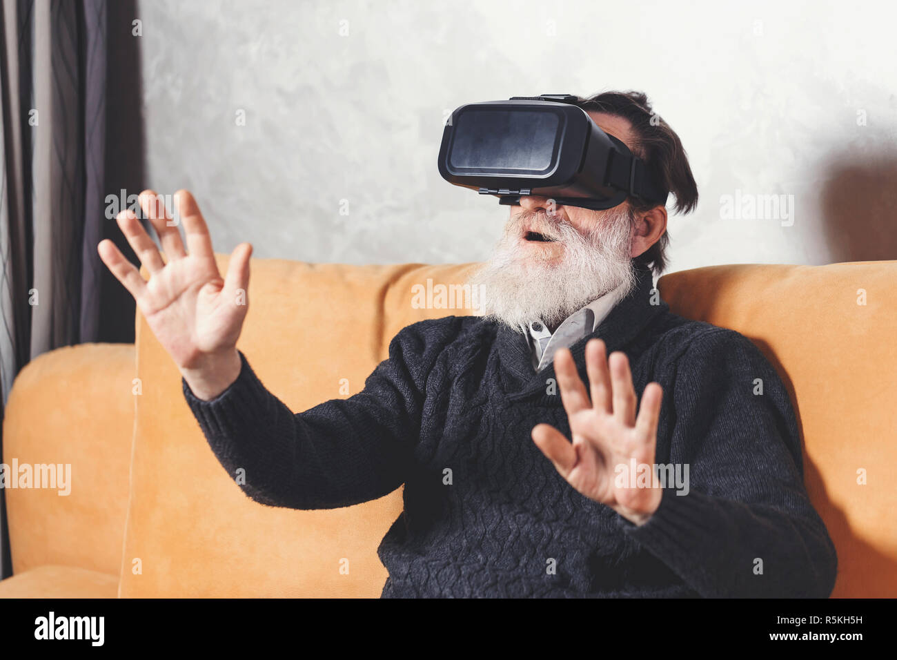 Überrascht älterer bärtiger Mann im grauen pullover Prüfung VR-Gerät während der Sitzung auf dem gelben Sofa im Wohnzimmer, zukünftige Technologie Konzept Stockfoto