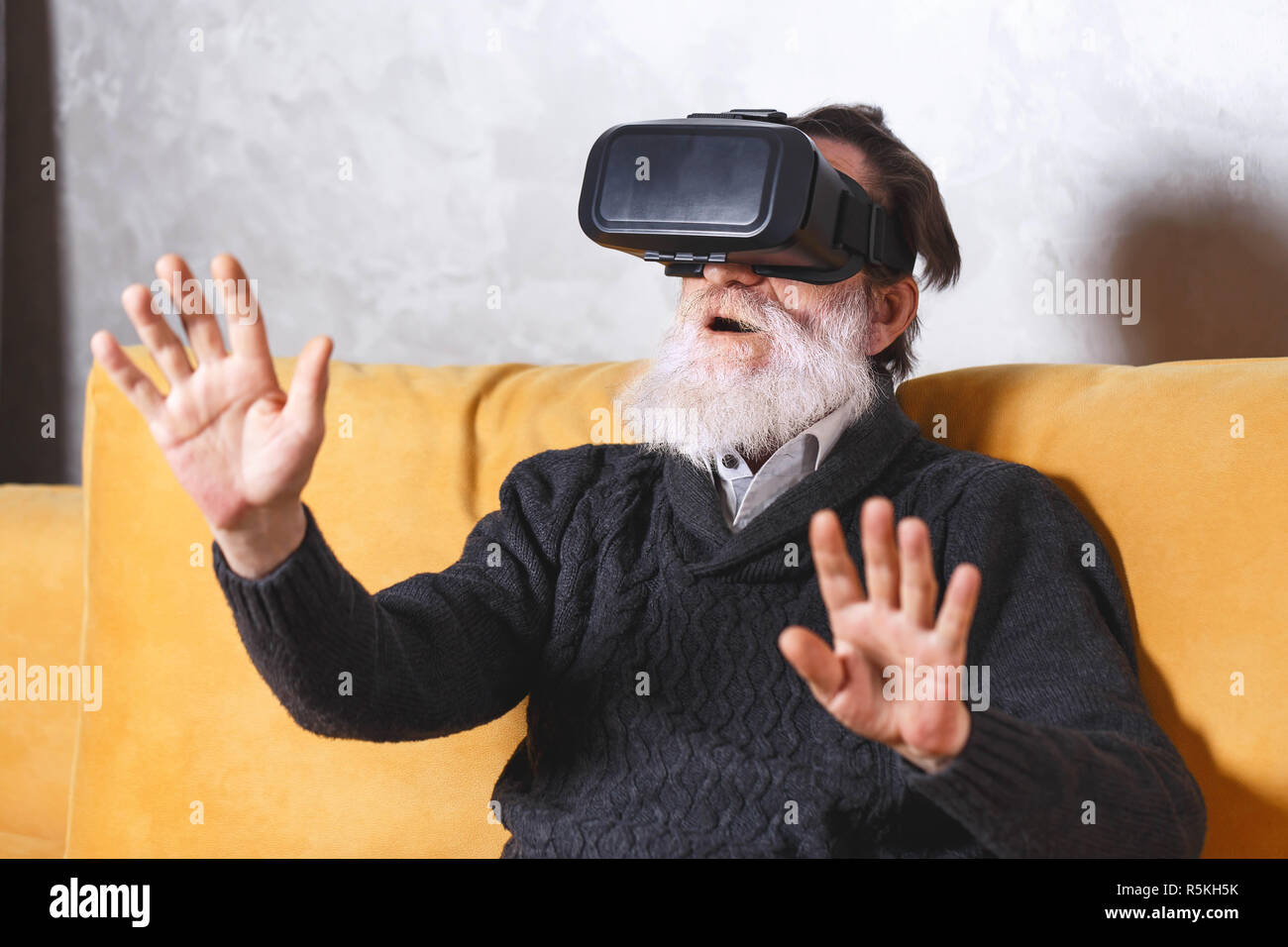 Überrascht älterer bärtiger Mann im grauen pullover Gestik, die während der Prüfung VR-Gerät, er saß auf dem gelben Sofa im Wohnzimmer, zukünftige Technologie Konzept Stockfoto