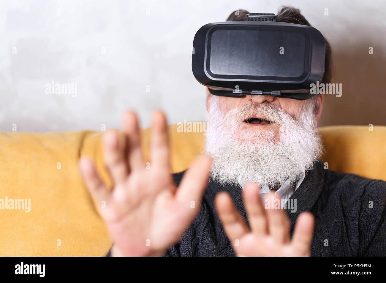 Überrascht älterer bärtiger Mann im grauen pullover etwas Berühren mit den Händen während der Prüfung VR-Gerät, er saß auf dem gelben Sofa im Wohnzimmer, zukünftige Technologie Konzept Stockfoto