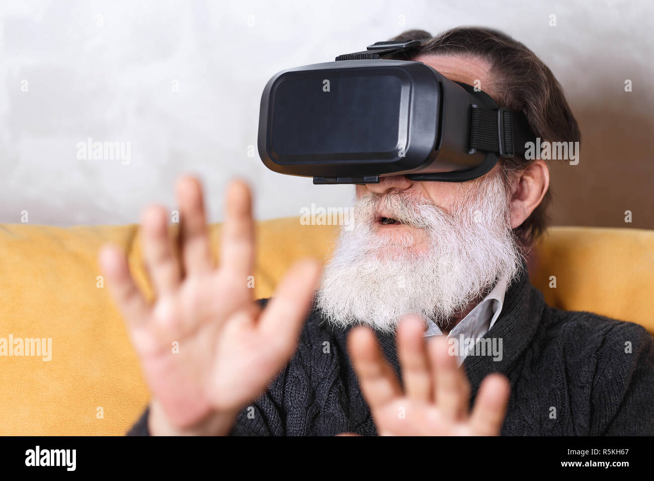 Überrascht älterer bärtiger Mann im grauen pullover Prüfung VR-Gerät, er saß auf dem gelben Sofa im Wohnzimmer, zukünftige Technologie Konzept Stockfoto