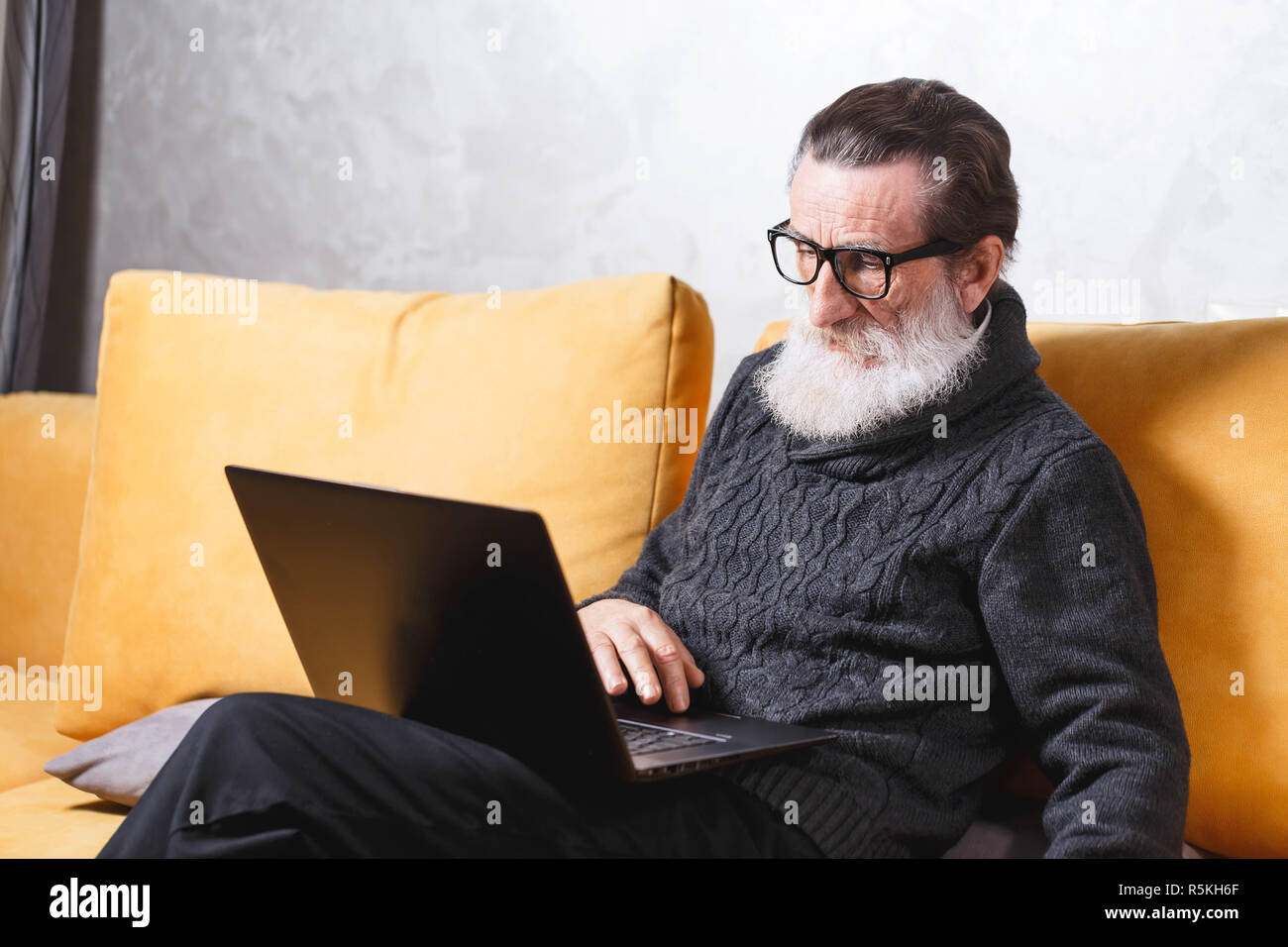 Gut aussehender älterer bärtiger Mann in Gläsern und grauen pullover sitzt mit Laptop auf dem gelben Sofa im Wohnzimmer, moderne Technologie, Kommunikation Konzept Stockfoto