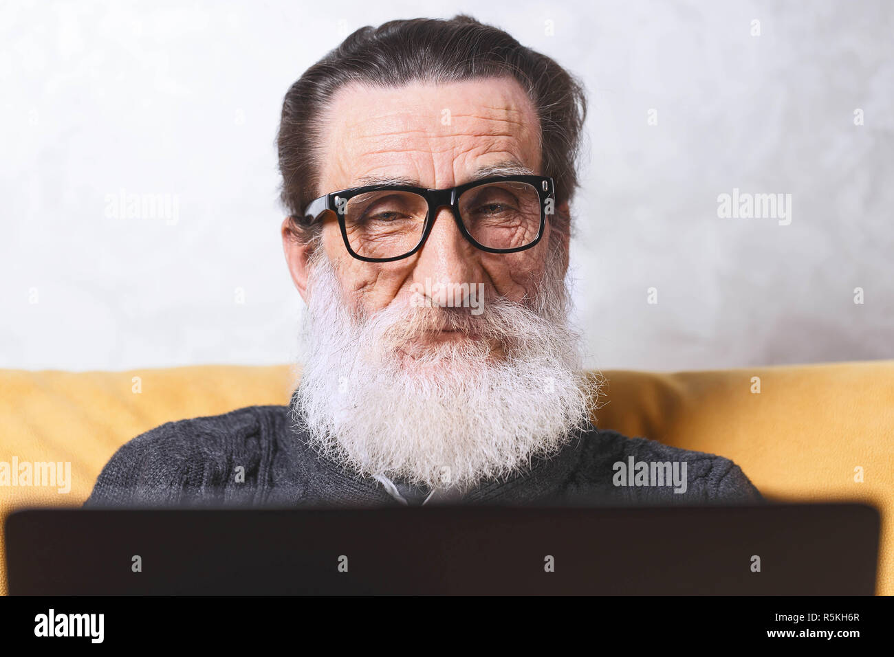 Fröhlichen älteren bärtigen Mann in Gläsern und grauen pullover auf Facebook Chat auf seinem Laptop sitzen auf dem gelben Sofa im Wohnzimmer, moderne Technologie, Kommunikation Konzept Stockfoto