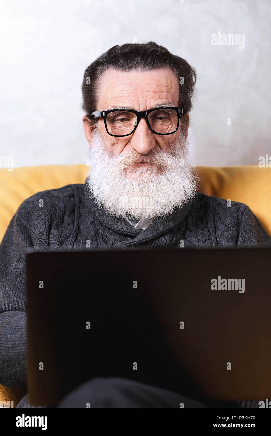 Fröhlichen älteren bärtigen Mann in Gläsern und Grau mit Laptop während der Sitzung auf dem gelben Sofa im Wohnzimmer pullover, moderne Technik, Kommunikation Konzept Stockfoto