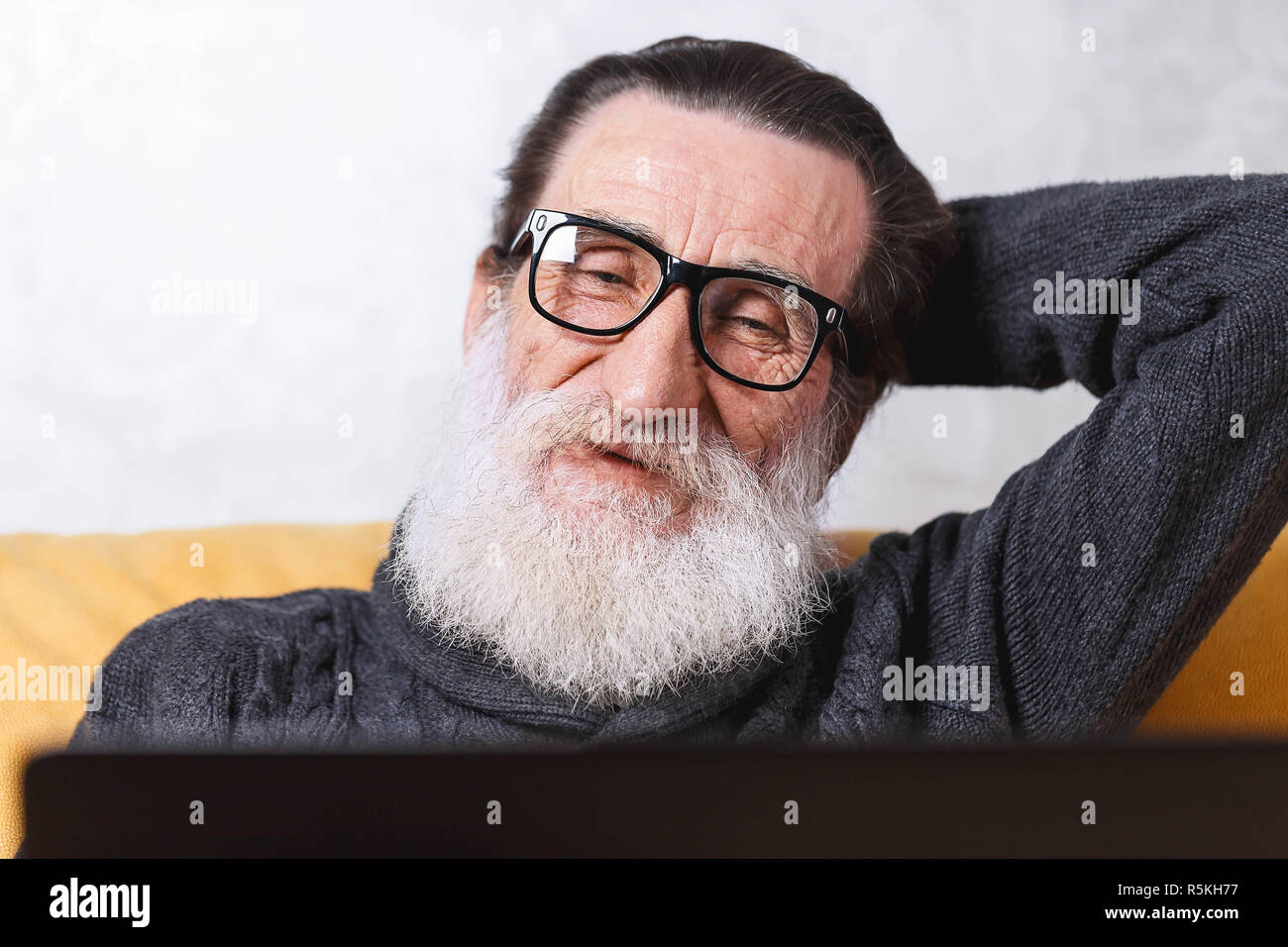 Fröhlichen älteren bärtigen Mann in Gläsern und grauen pullover Ansehen guter Film auf seinem Laptop sitzen auf dem gelben Sofa im Wohnzimmer, moderne Technologie, Kommunikation Konzept Stockfoto