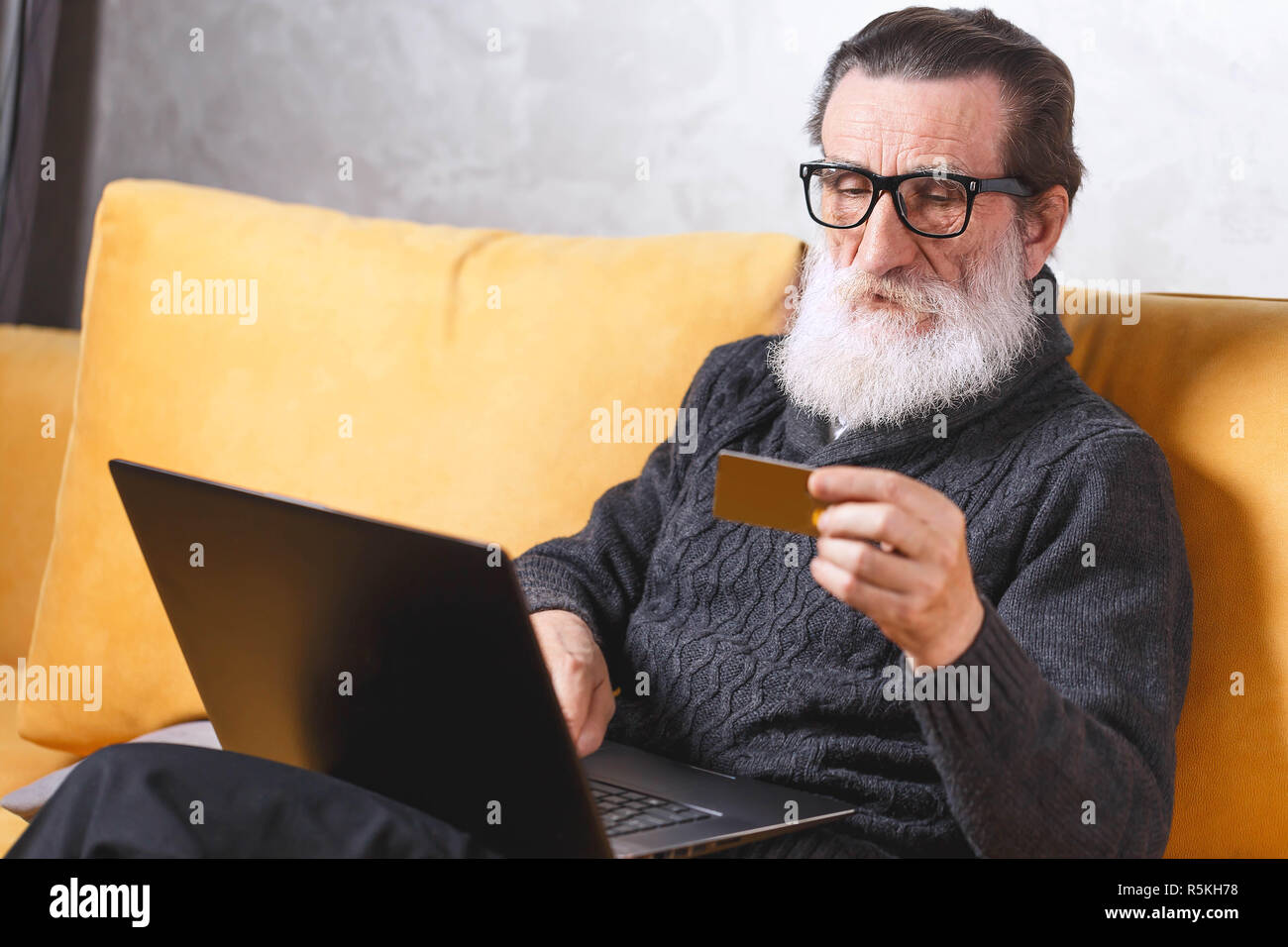 Fröhlichen älteren bärtigen Mann in Gläsern und grauen pullover Einkauf mit Laptop und seine Kreditkarte beim Sitzen auf dem gelben Sofa im Wohnzimmer, moderne Technologie, Kommunikation Konzept Stockfoto