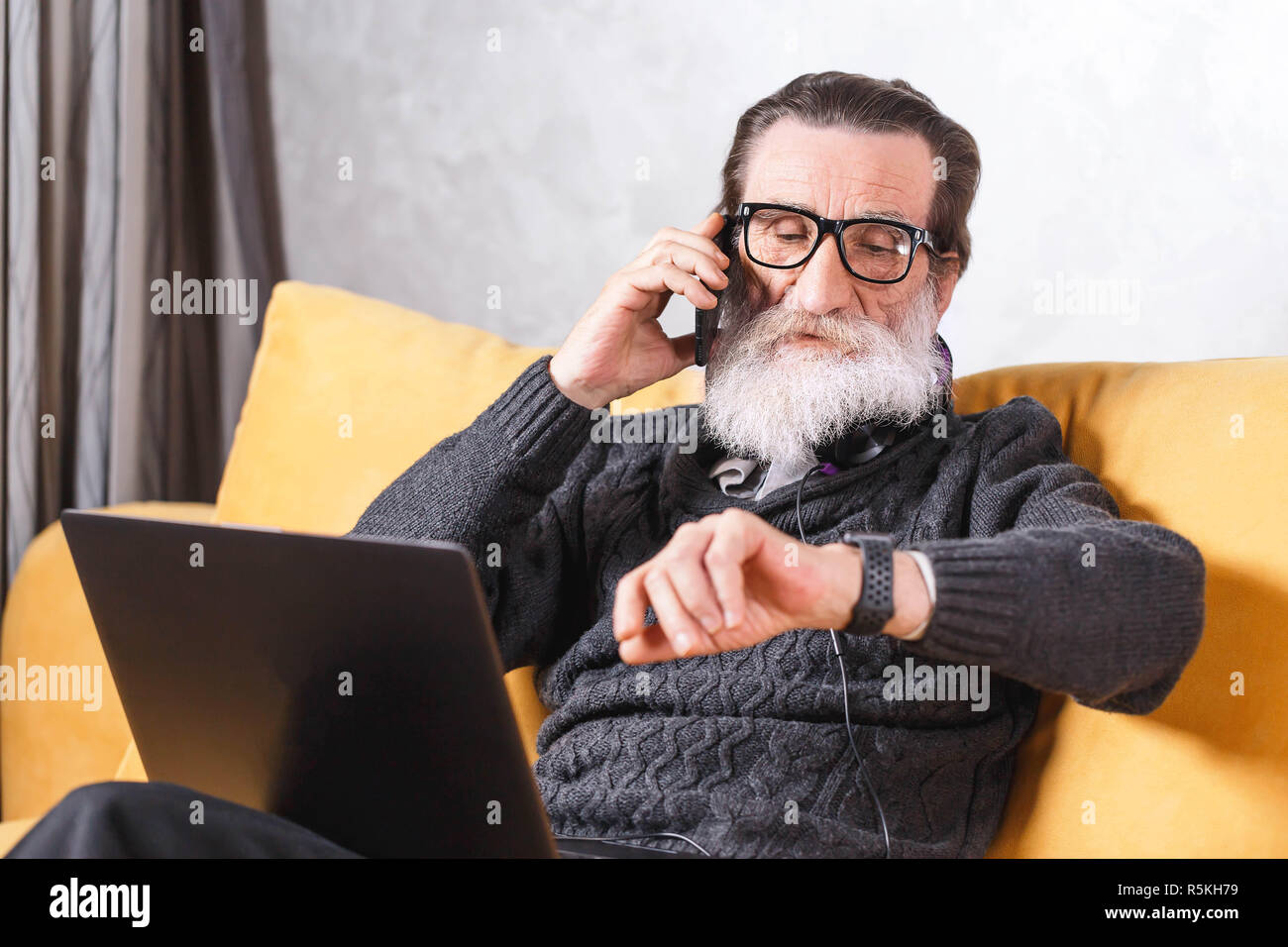 Zeitgenössische älterer bärtiger Mann in Gläsern und grauen pullover Plaudern auf dem Smartphone und schauen mal in seinem Apple beobachten, während das Sitzen auf dem gelben Sofa im Wohnzimmer, moderne Technologie, Kommunikation Konzept Stockfoto