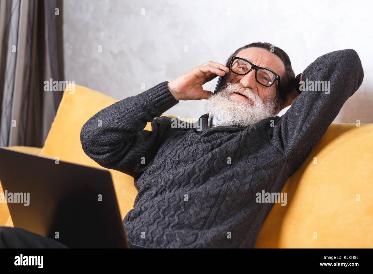 Zeitgenössische älterer bärtiger Mann in Gläsern und grauen pullover Entspannen und Plaudern auf dem Smartphone mit seinem Freund beim Sitzen auf dem gelben Sofa im Wohnzimmer, moderne Technologie, Kommunikation Konzept Stockfoto