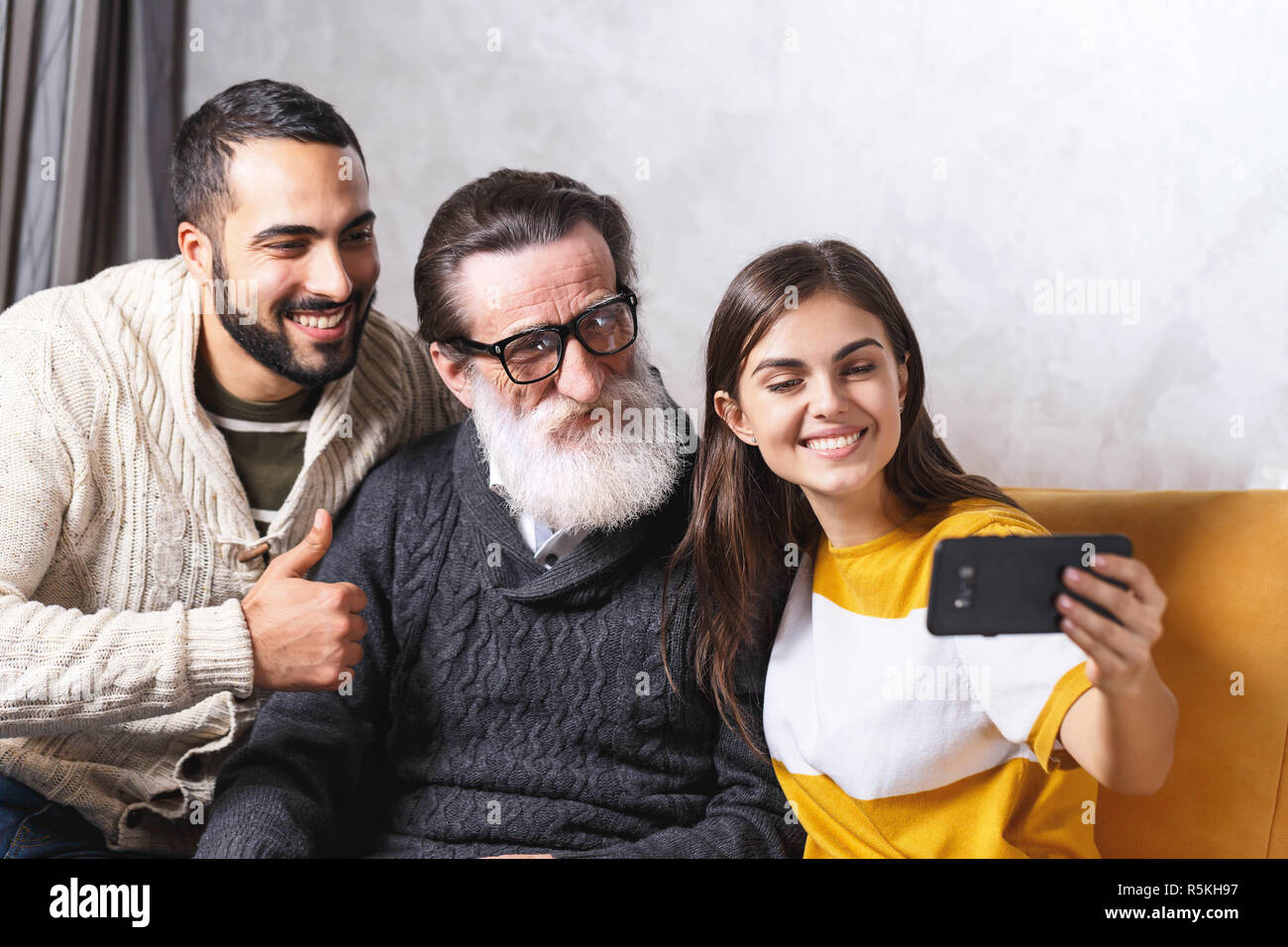 Brunette bärtigen Sohn und Brunette langhaarigen Tochter lächelnd und mit selfie sie älteren bärtigen Vater in Gläser beim Sitzen auf dem gelben Sofa im Wohnzimmer, moderne Technologie, Kommunikation Konzept Stockfoto
