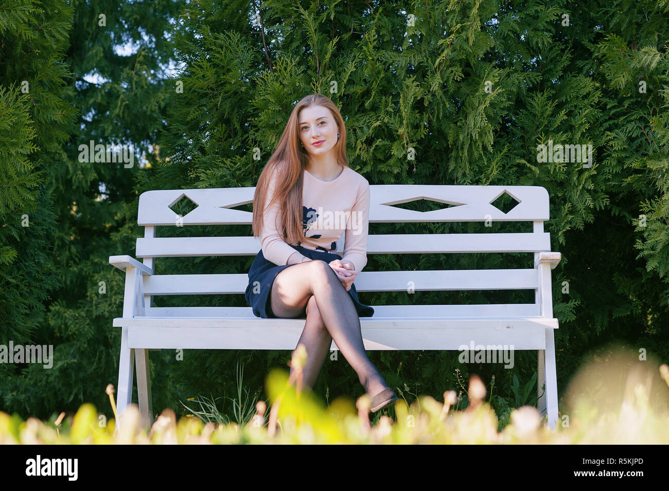 Ein nettes Mädchen mit langen roten Haaren, sitzt auf einer weißen Bank vor dem Hintergrund eines Gartens mit Lebensbäume und lächelt. Porträt im freien Stockfoto