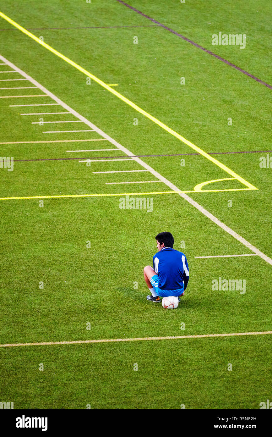 Unglücklich Teenager aus der Fußball-Mannschaft ausgeschlossen sitzt auf den Ball an der Ecke der grünen Fußballplatz. Stockfoto