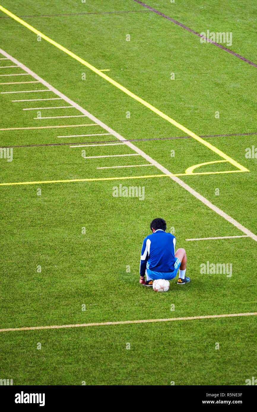 Unglücklich Teenager aus der Fußball-Mannschaft ausgeschlossen sitzt auf den Ball an der Ecke der grünen Fußballplatz. Stockfoto