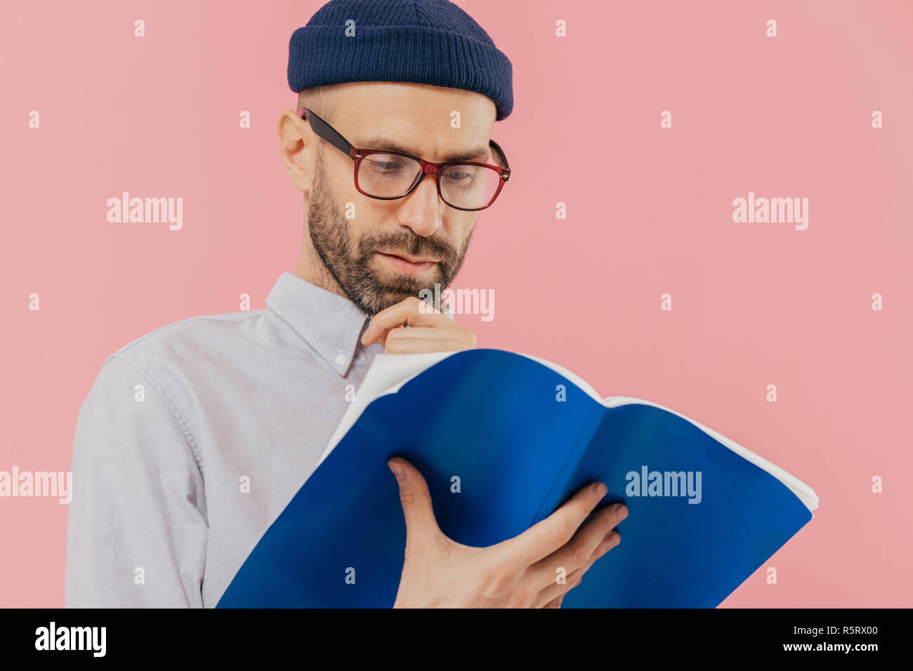 Nahaufnahme von ernster Mann Kinn hält, hat dunkle Haare, trägt eine Brille für eine gute Vision, trägt Buch geöffnet, lernt Informationen, steht gegen Rosa Stockfoto