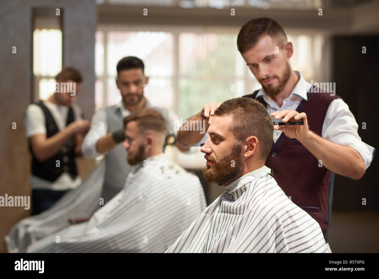 Seitenansicht des Prozesses der Hairstyling in Friseur. Junge Bartgeier im Stuhl sitzen, im Spiegel zu betrachten und sich die Haare schneiden. Professionelle Friseur in Uniform, Schere und Rasiermesser. Stockfoto