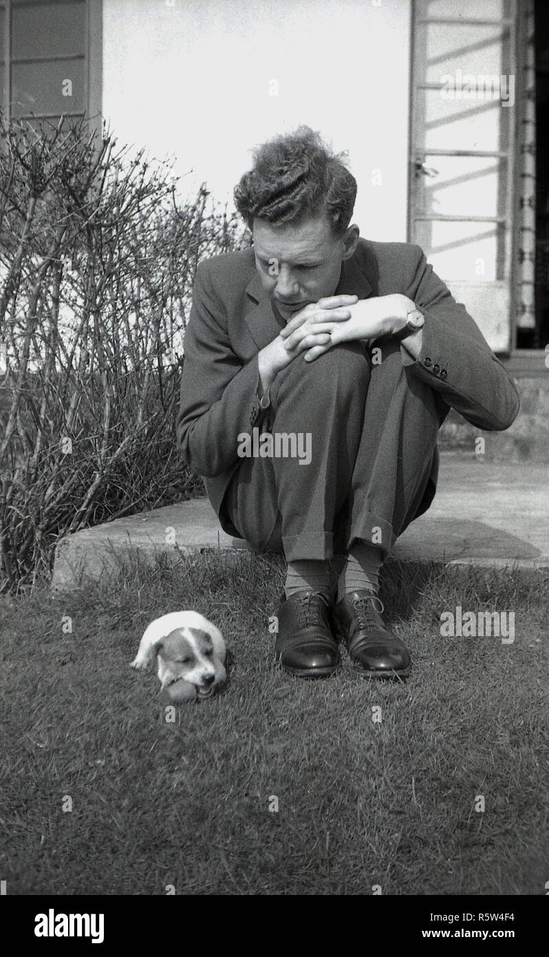 1950 s, historischen, einen geeigneten Mann sitzt auf einem Schritt in einem Garten neben einem kleinen Jack Russell Terrier Hund. Der Jack Russell Terrier wurde ursprünglich als working Terrier zu jagen Füchse gezüchtet und ist eine lebendige, unabhängige und kluge kleinen Hund. Stockfoto