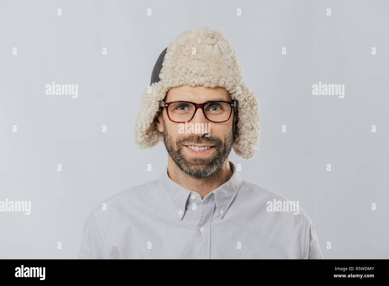 Headshot fröhliche stattlichen Kaukasischen Kerl trägt warme Mütze mit Ohrenschützern, Brillen und weißes Hemd, sieht direkt an der Kamera, bereit für übertreffen Stockfoto