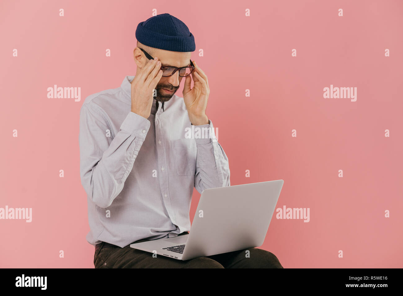 Nachdenklich ernsten Kursteilnehmer hält die Hände auf spectalces, hat online Gespräch via Notebook Computer, Uhren webinar, trägt Hut und weissen Hemd Stockfoto