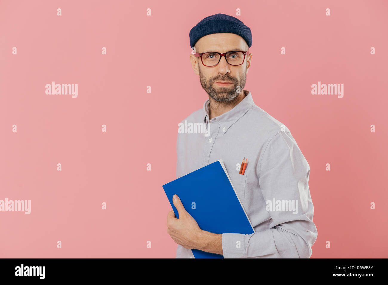 Stattliche männliche Designer trägt stilvolle Kleidung, hat zwei Stifte in der Tasche von Hemd, hält Blau notepad für Sketche und über Rosa backgroun isoliert Stockfoto