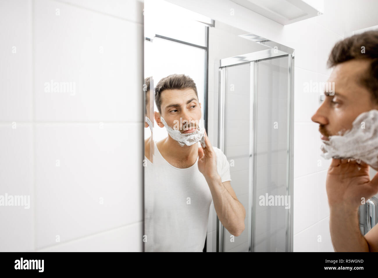 Schöner Mann im weißen T-Shirt shawing seinen Bart mit Klinge und Schaum im Badezimmer Stockfoto