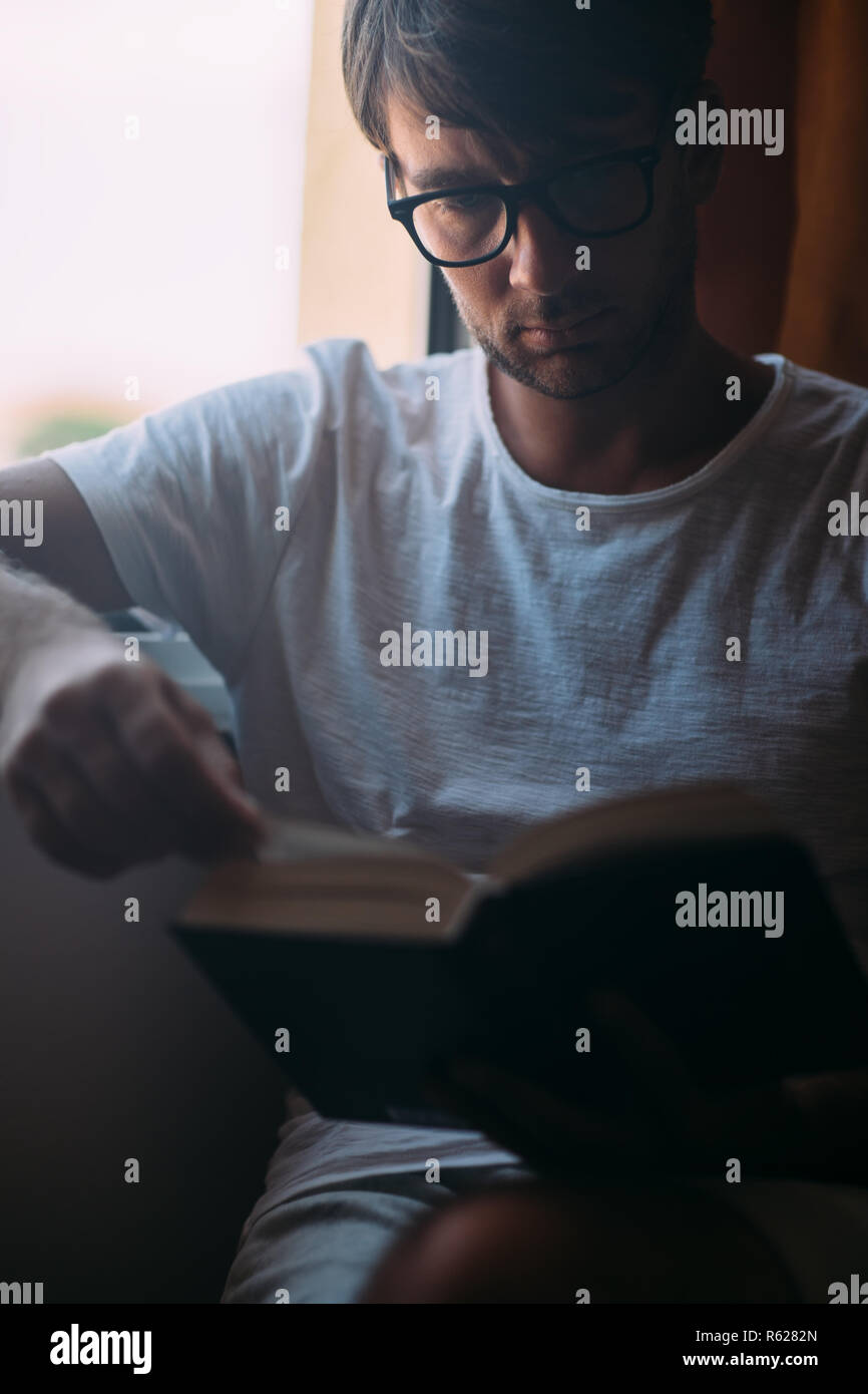 Jungen gutaussehenden Mann in Brillen lesen Buch sitzen in der Nähe der Fenster. Wochenende Konzept Stockfoto
