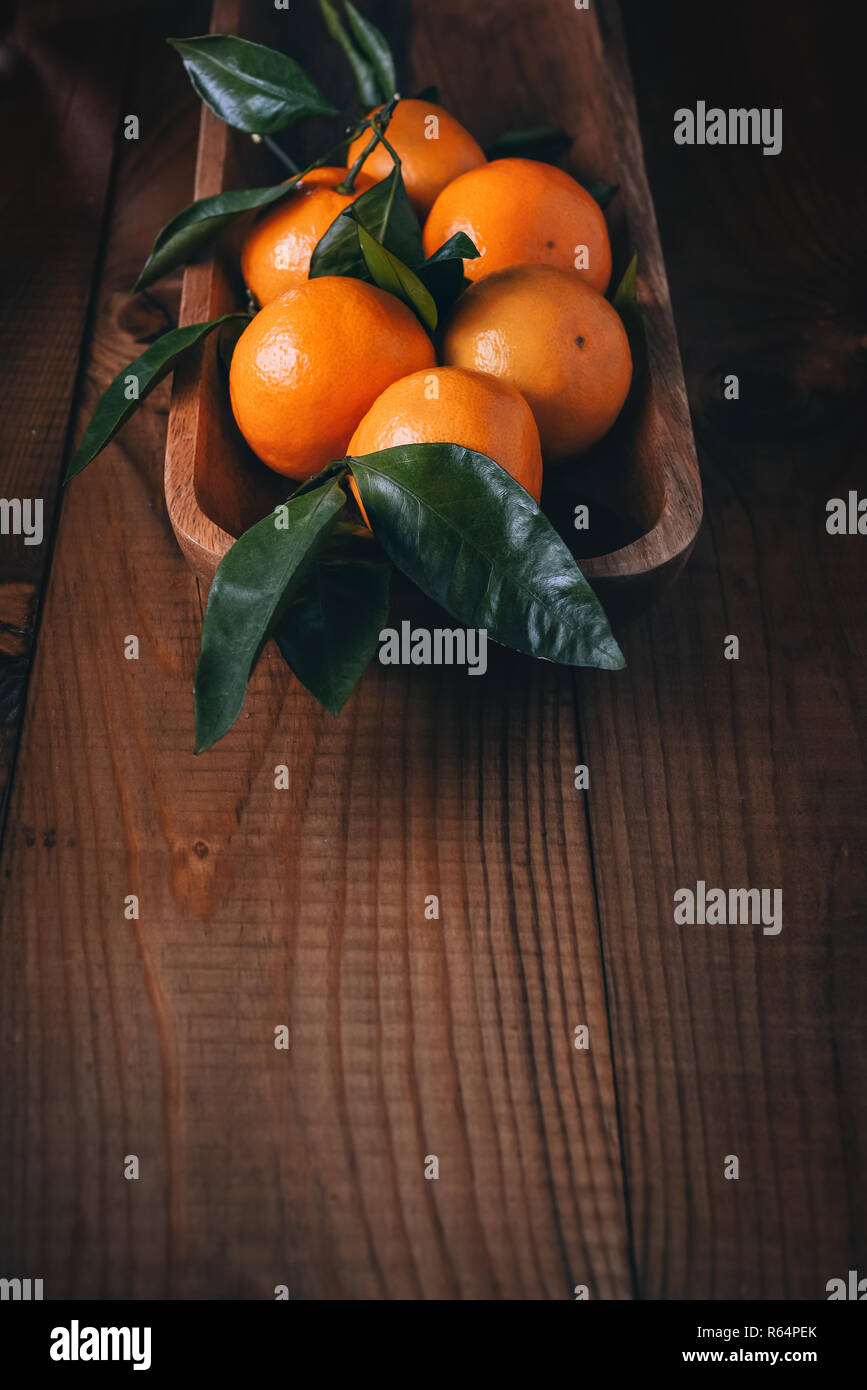 Viele Mandarinen mit grünen Blättern in einem hölzernen Schale auf einem dunklen Holztisch. Vintage Stockfoto
