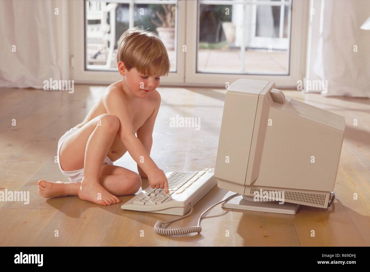 Porträt, Innenraum, 6-jaehriger blonder Junge nur mit Unterhose bekleidet sitzt mit dem Holzboden und spielt am Computer Stockfoto