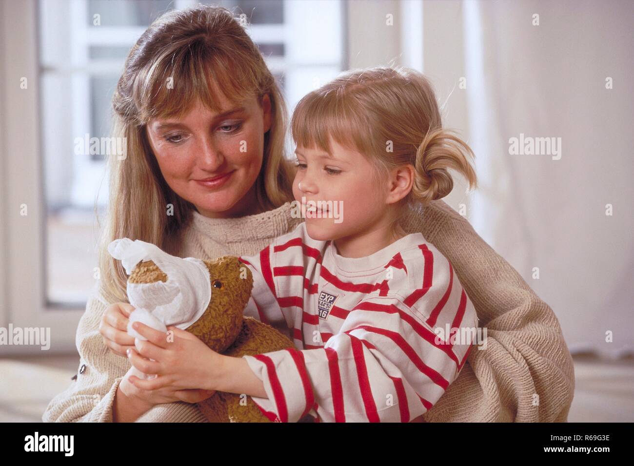 Porträt, Innenraum, Hölle gekleidete blonde Frau und ihre 6-jährige blonde Tochter mit rot- weiss gestreiftem T-Shirt Artikel mit dem Boden und spielen mit einem Teddy Stockfoto