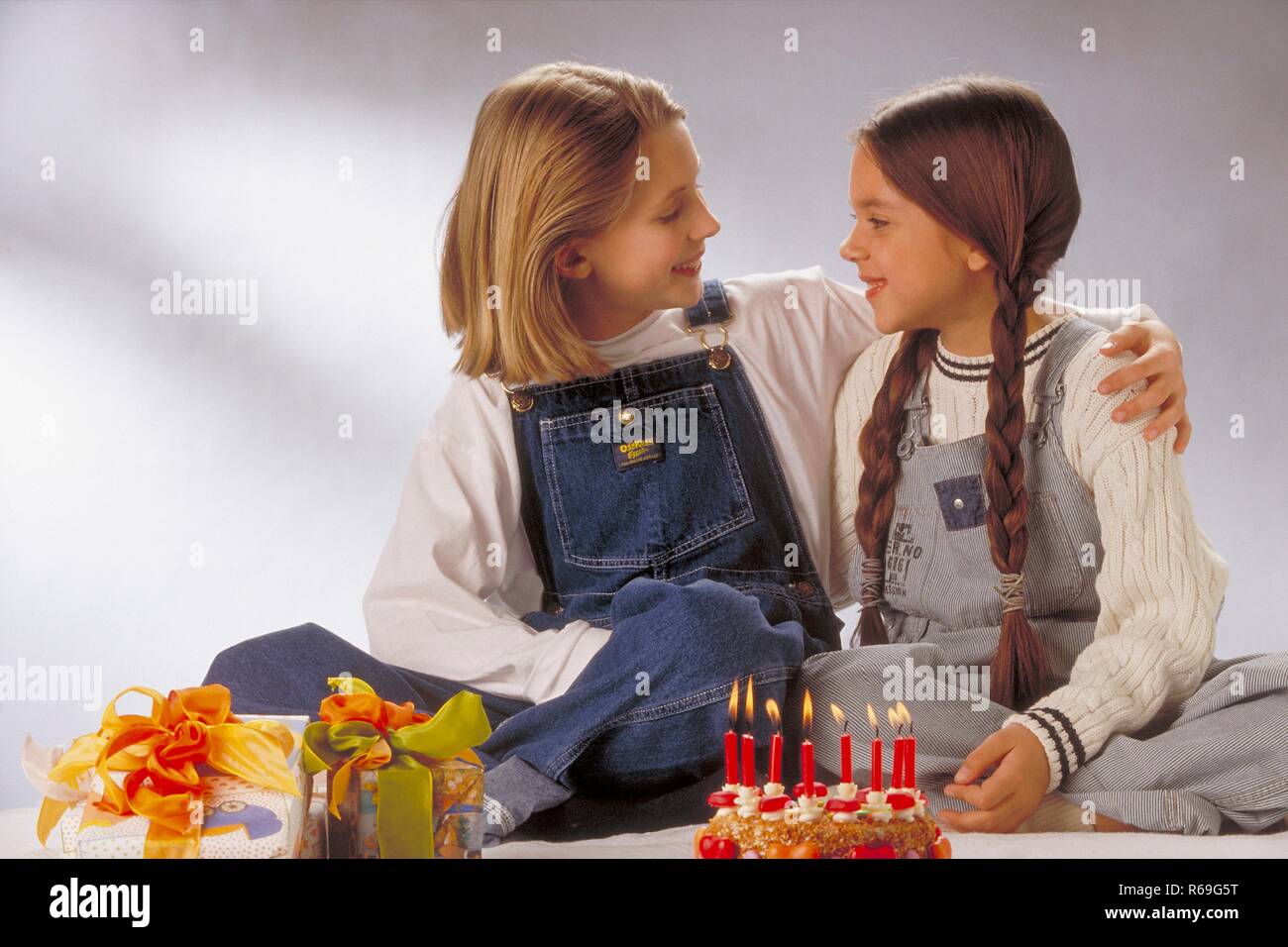 Halbfigur, Innenraum, 2 Maedchen bekleidet mit Jeanslatzhosen Artikel mit einen Geburtstagskuchen mit Herzen und 8 brennenden Kerzen auf dem Boden Stockfoto