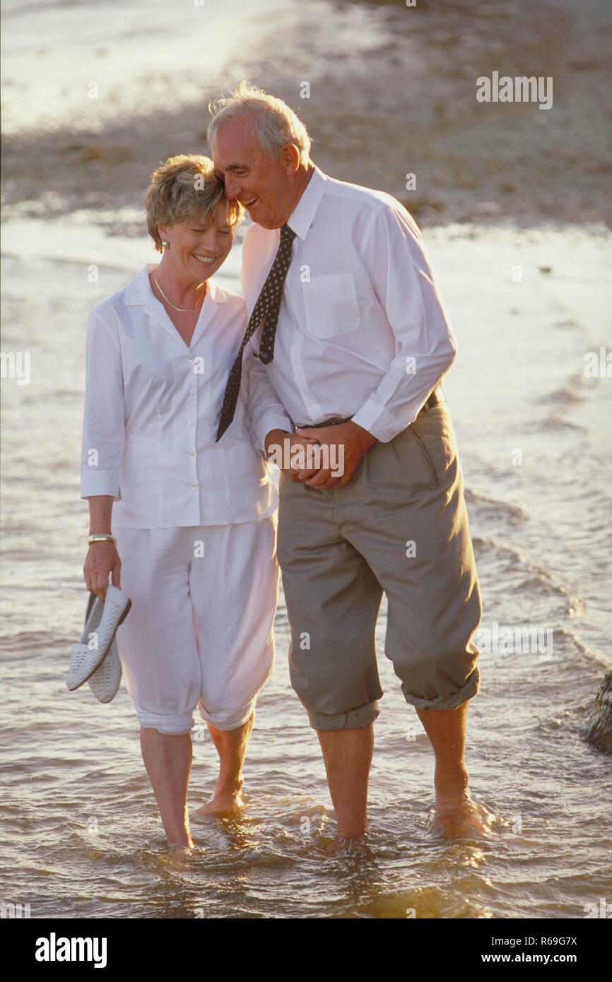 Ganzfigur, Seniorenpaar, weiss gekleidete Frau und Mann mit weissem Pullover, Hofgarten mit den Schuhen in der Hand barfuss Hand in Hand durchs seichte Wasser Stockfoto