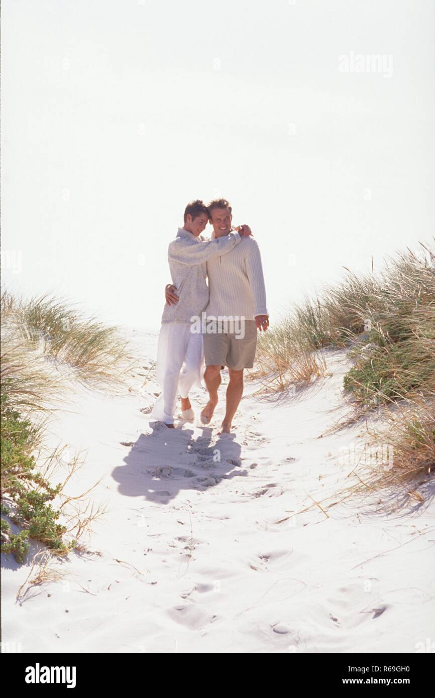 Strandszene, Ganzfigur, Mann und Frau mit kurzen braunen Haaren bekleidet, mit naturfarbenen Wollpullovern und hellen Hosen gehen Arm in Arm durch die Duenen Stockfoto