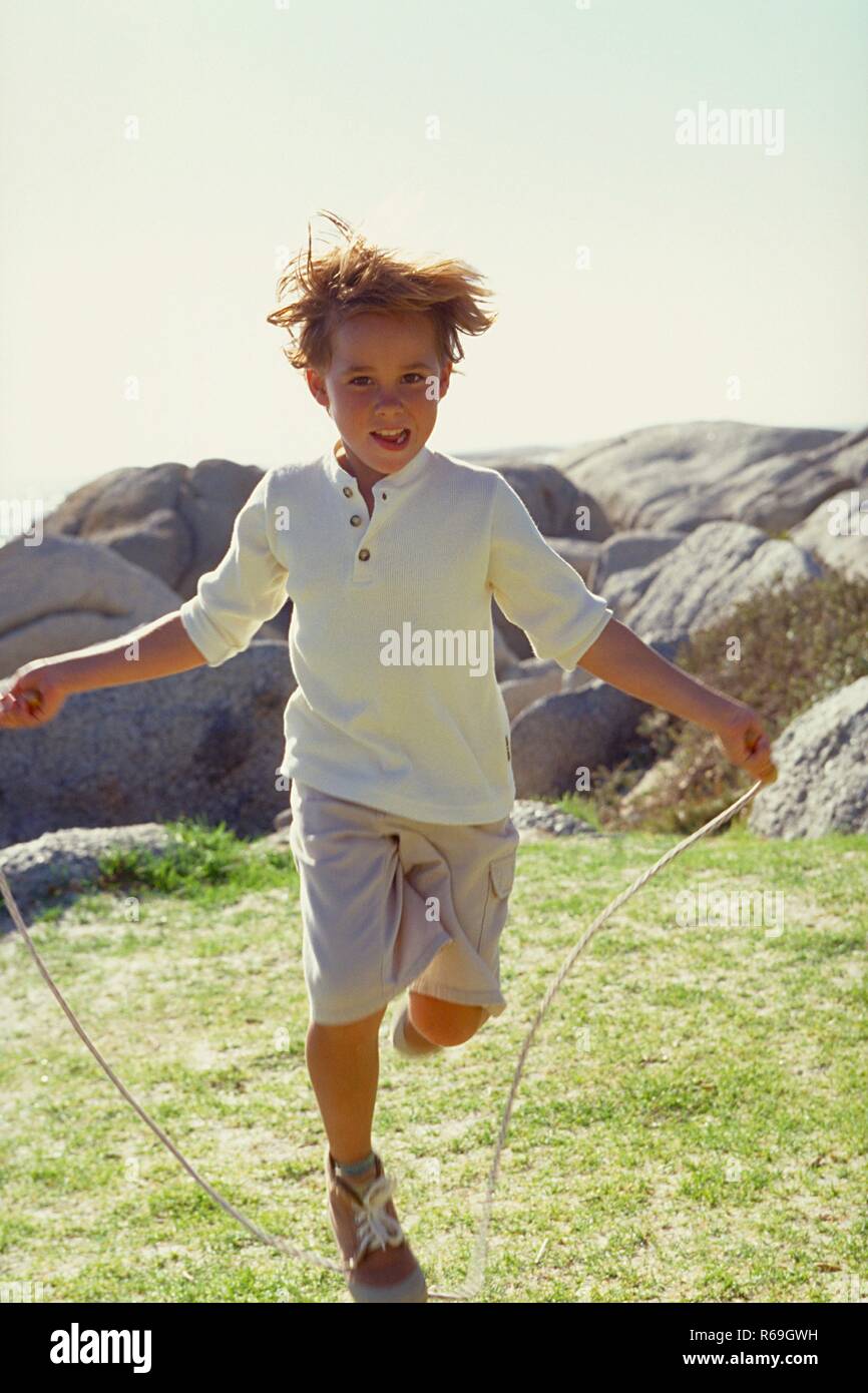 Ganzfigur, 10 Jahre alter blonder Junge, bekleidet mit heller knielanger Schlauch, weissem Shirt und beigen Turnschuhen springt Seilchen in einer Wiese vor Felsen Stockfoto