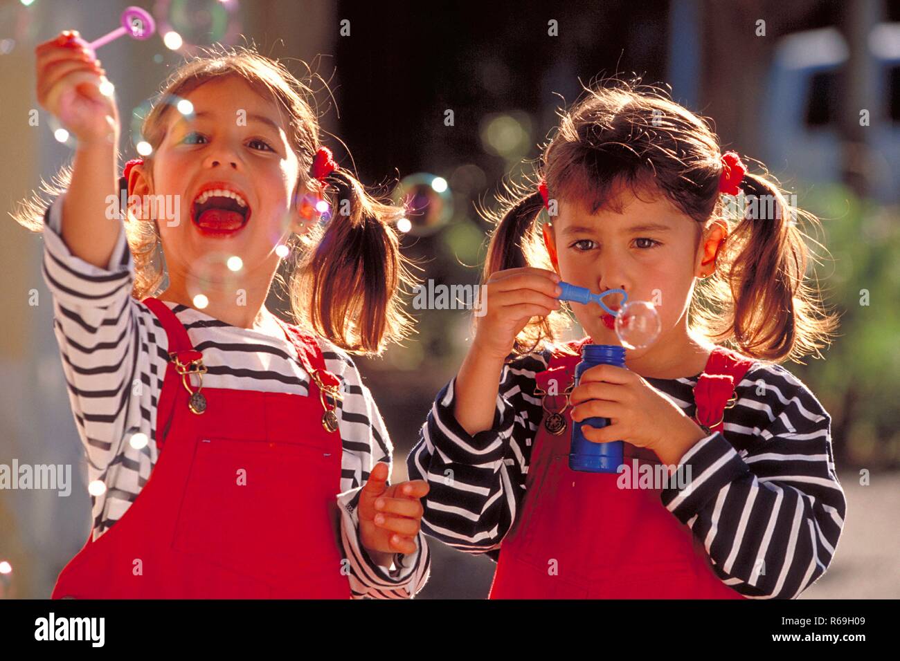 Porträt, Halbfigur, zwei 5 Jahre alte Zwillings-Maedchen mit kurzen Zoepfen bekleidet mit gestreiftem T-Shirt und roter Latzhose halten kleine Flaschen in der Hand und Seifenblasen blasen Stockfoto