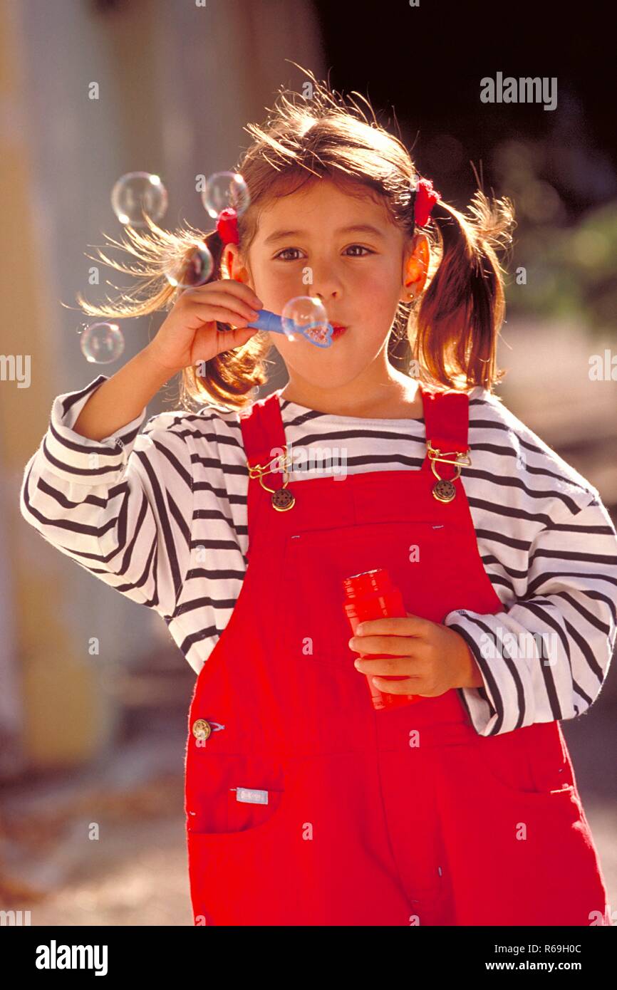 Porträt, Halbfigur, 5 Jahre altes Maedchen mit kurzen Zoepfen bekleidet mit gestreiftem T-Shirt und roter Latzhose haelt eine rote Flasche in der Hand und pustet Seifenblasen Stockfoto