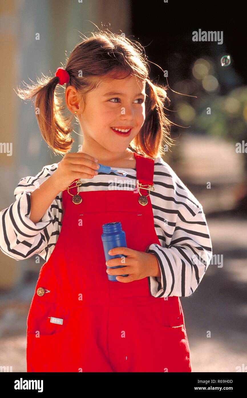 Porträt, Halbfigur, 5 Jahre altes Maedchen mit kurzen Zoepfen bekleidet mit gestreiftem T-Shirt und roter Latzhose haelt eine blaue Flasche Seifenblasen in der Hand Stockfoto
