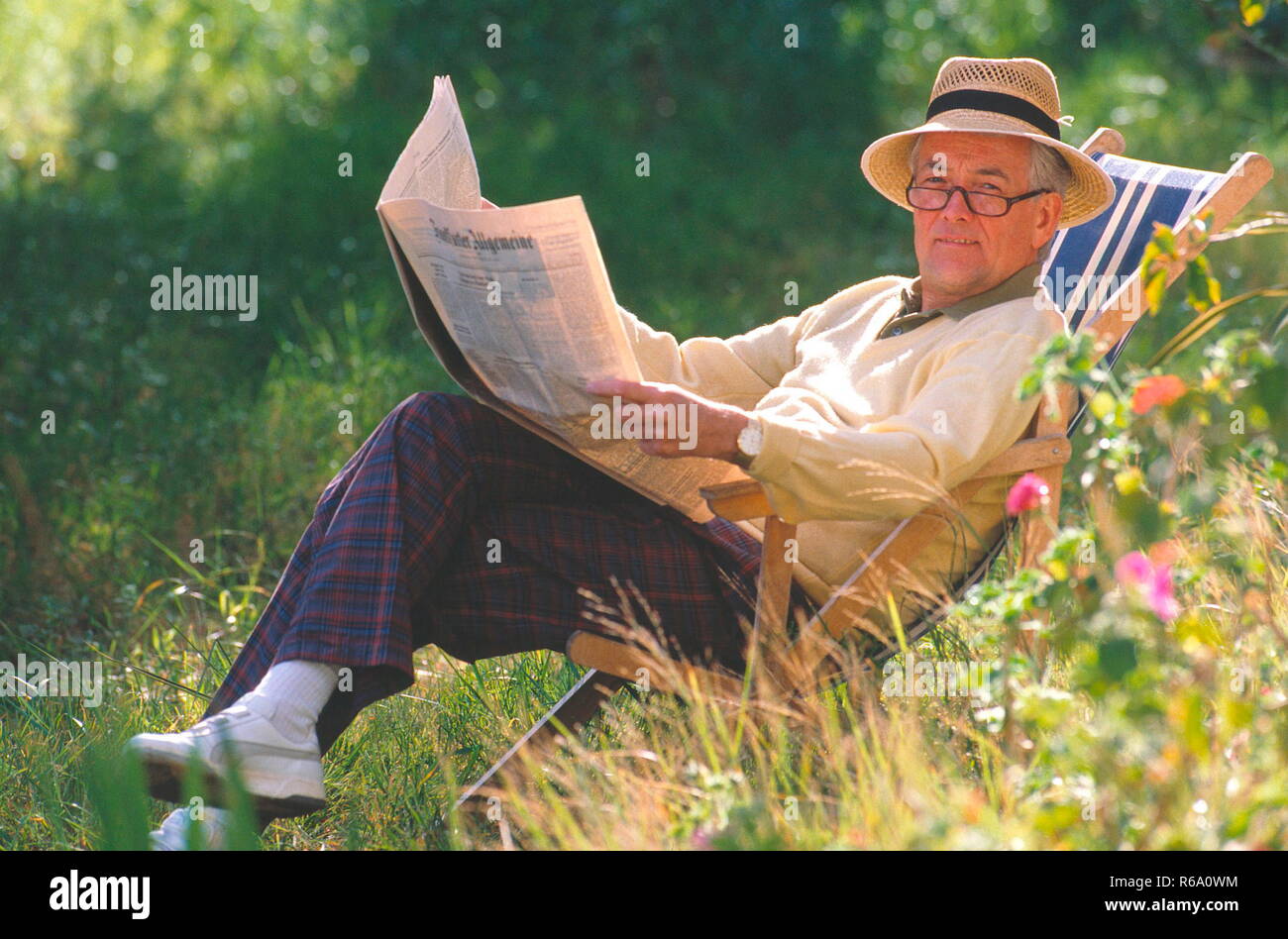 Portrait, Senior in Freizeitkleidung mit Brille und Strohhut sitzt im Garten im Liegestuhl und liest seine Zeitung Stockfoto
