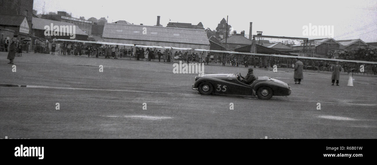 1950, historische, an einem Industriestandort, eine ungewöhnliche offenen Sportwagen entlang gefahren wird, von den Zuschauern beobachtet. Stockfoto