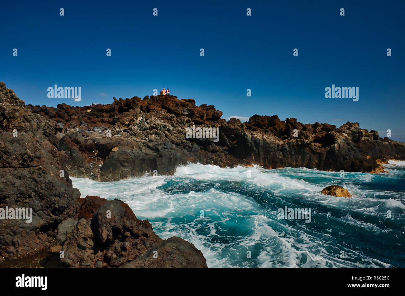 Shoreline, Los Cancajos. La Palma. Die Lava Rock Küsten Relief, die typisch für die Insel sind. Zwei Touristen sitzen auf einer öffentlichen Bank hoch oben auf den Felsen, die Aussicht zu genießen. Es ist ein Sonniger Tag mit einem blauen Himmel. Stockfoto